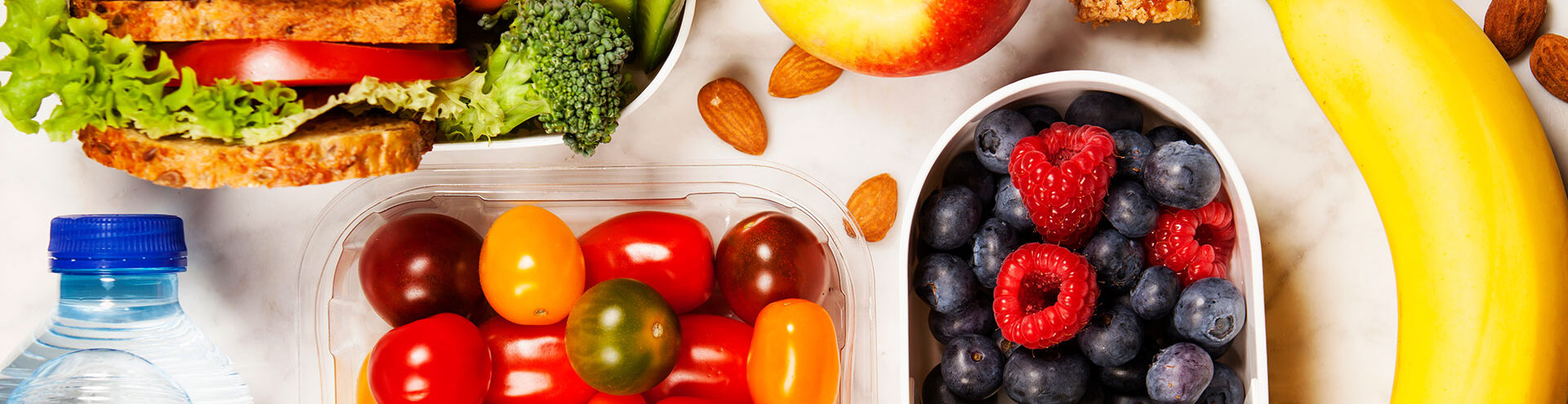 A sandwich, apple, almonds, banana, grape tomatoes, blueberries, raspberries, and a bottle of water are arranged on a white surface.