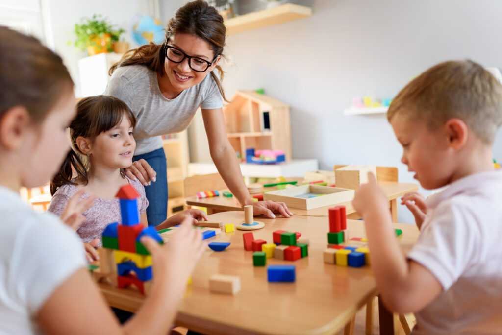 A teacher smiles and interacts with three young children playing with colorful building blocks at a wooden table in a bright classroom.