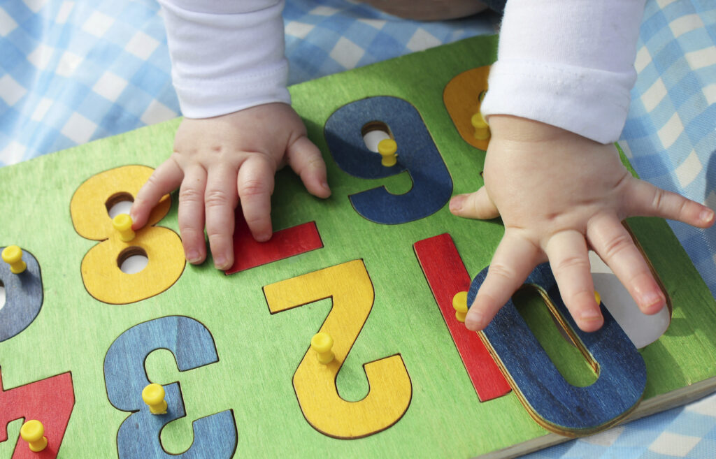 A baby’s hands touch colorful wooden puzzle numbers on a green board, with a blue and white checkered cloth underneath.