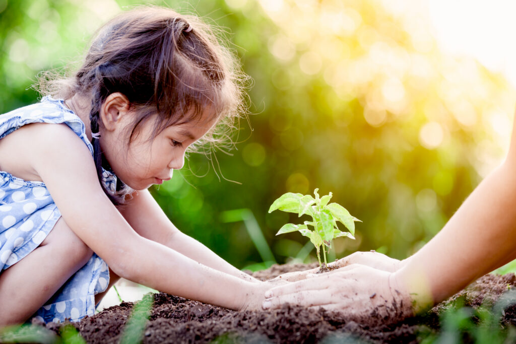 A young girl with brown hair, wearing a blue polka-dot dress, kneels on the ground and gently helps plant a small green seedling in the soil with another person's hands beside her. Sunlight shines in the background.