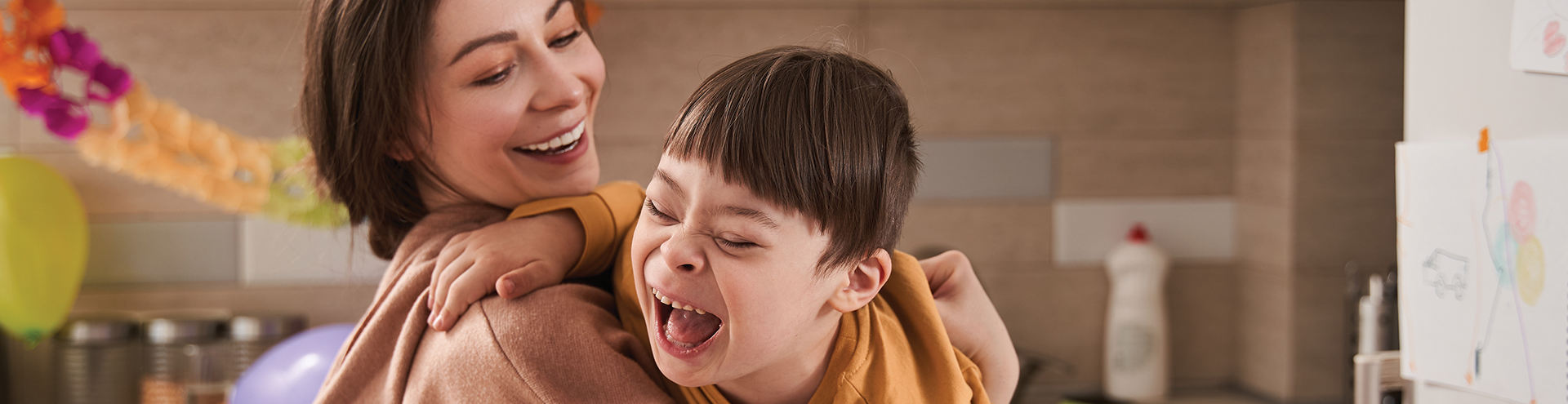 A woman hugs a laughing child with Down syndrome in a bright kitchen decorated with colorful streamers and balloons, creating a joyful and playful atmosphere.