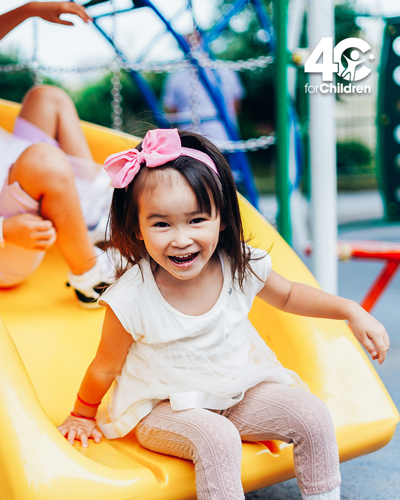A young girl with a pink bow in her hair smiles while sitting on a yellow playground slide, wearing a white dress. Another child is behind her.
