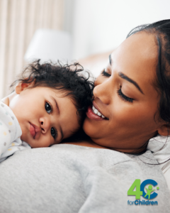 A woman smiles while holding a baby who is resting on her chest. The baby looks calm and cozy. In the corner is a logo that reads "40 for Children" with a green and blue color scheme.